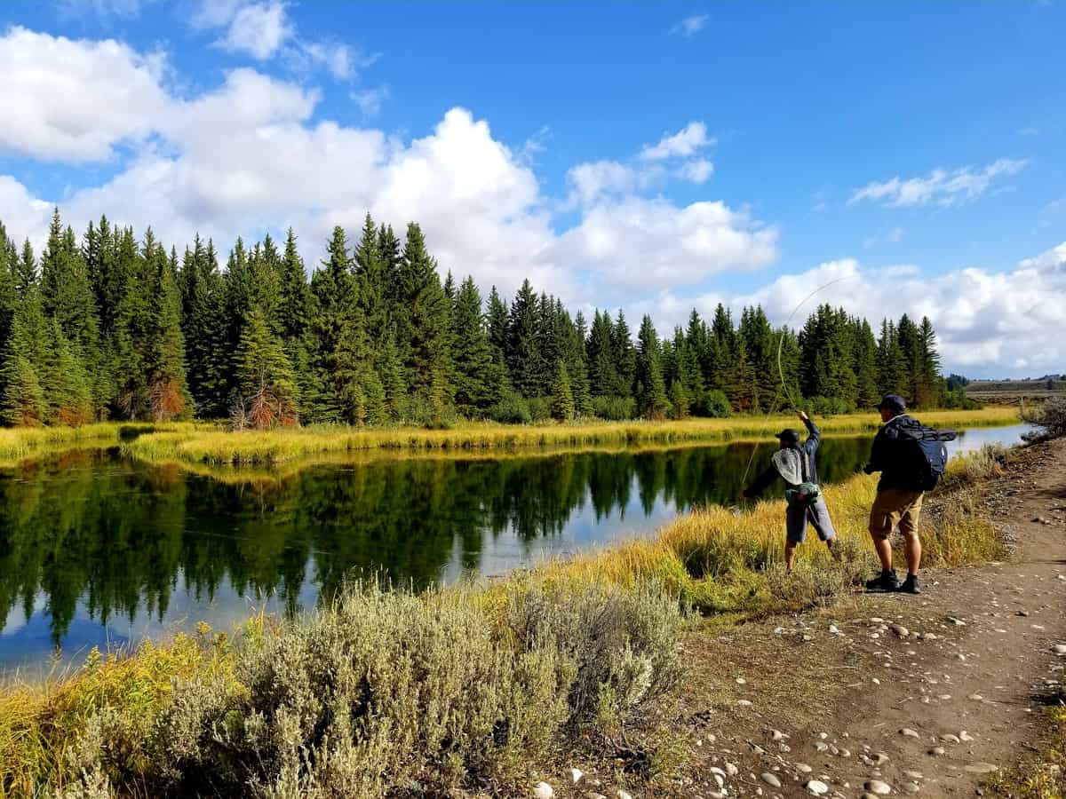 Two men fly fishing in a river with blue sky and lush greenery